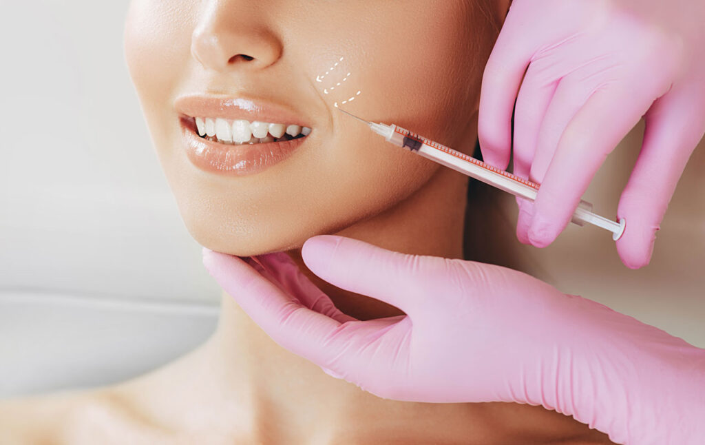 Clinician gives a fine facial filler injection near the cheek while supporting the patient's chin with gloved hands.