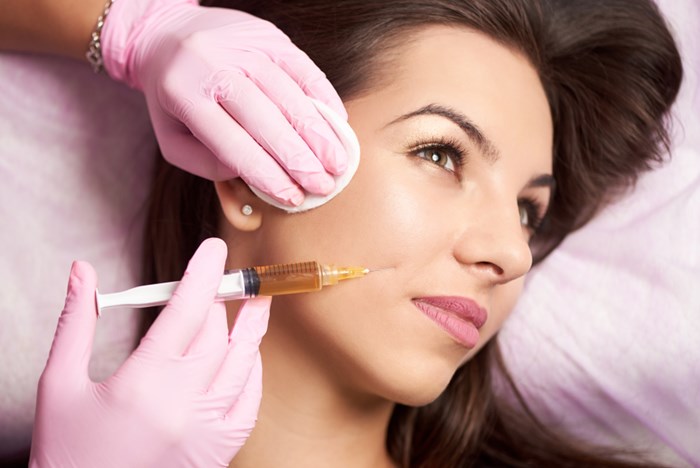 A clinician in pink gloves holds a syringe near a woman's cheek during treatment with an injectable facial filler.