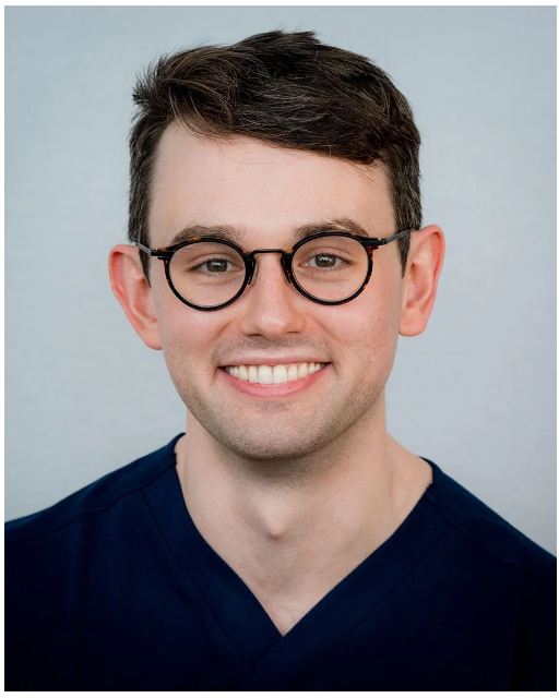 A smiling Dr Lachlan Byth wears round glasses and navy scrubs in a professional portrait for the Dermatology & Vein Institute Australia team profile.