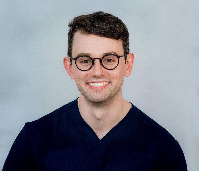 Portrait of Dr Lachlan Byth, a smiling dermatologist wearing navy medical scrubs and round glasses